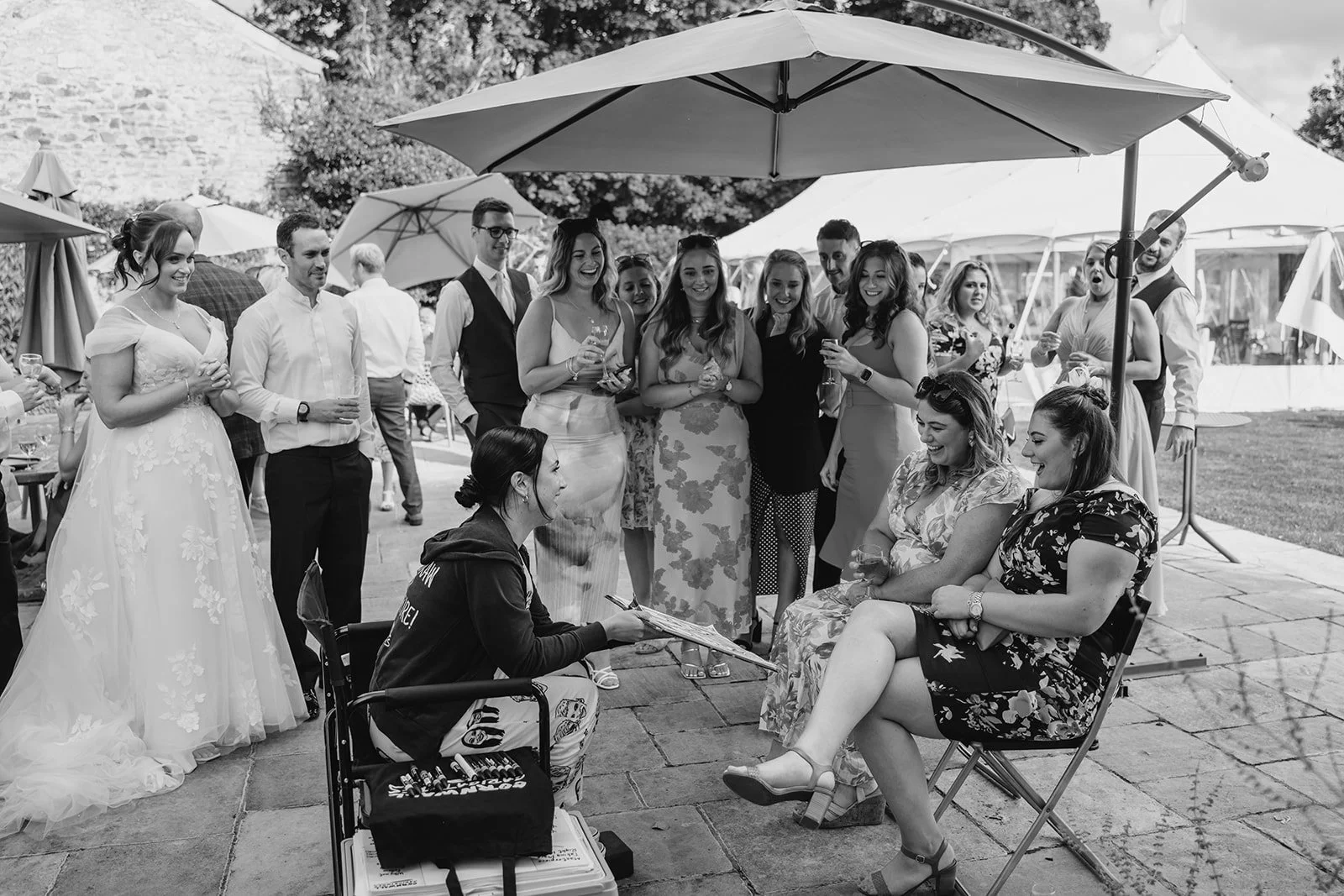 Group of people at an outdoor wedding reception, with some women in dresses and men in shirts, gathered under umbrellas on a patio, smiling and talking, with two women sitting in chairs getting their caricatures drawn and others standing around them.