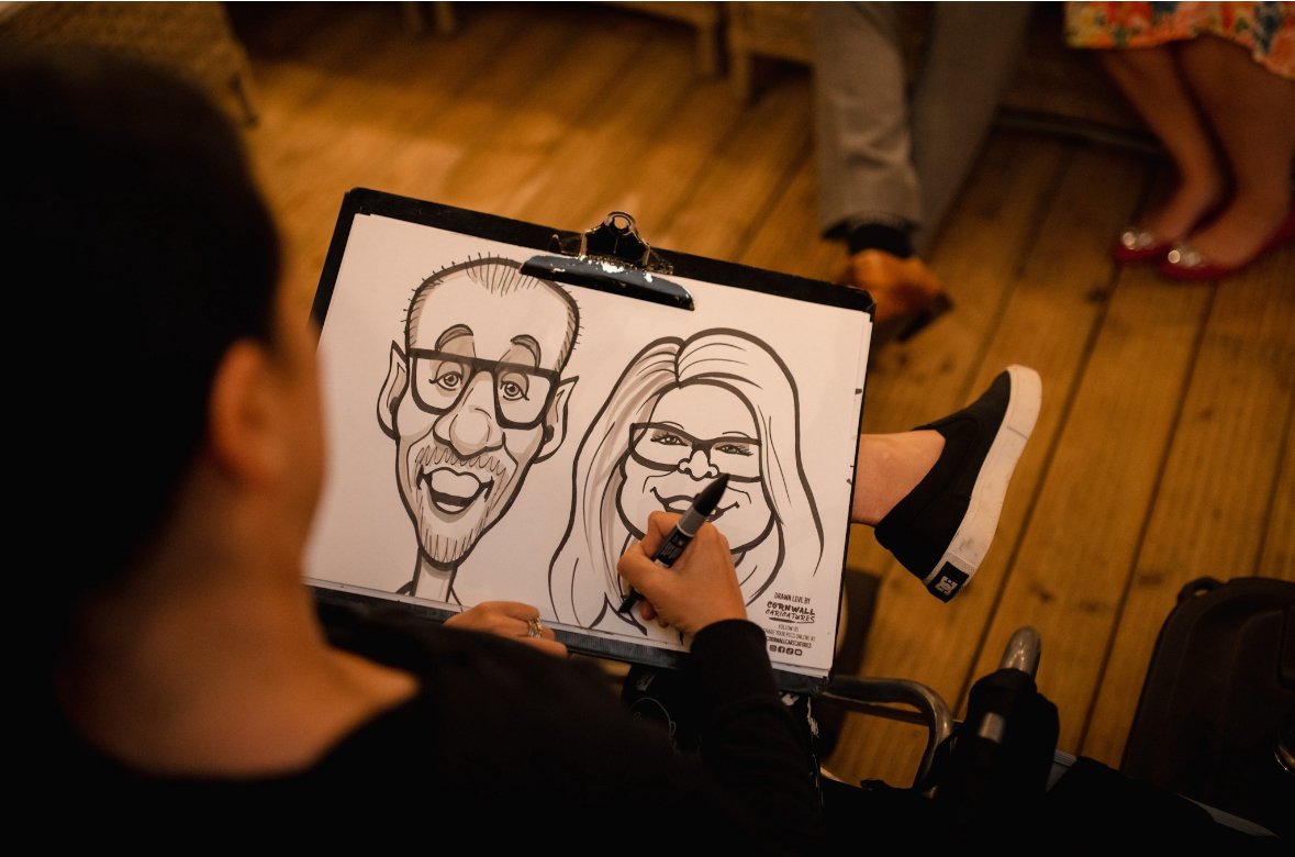 A person is drawing cartoon-style caricatures of a man and a woman on a clipboard during an event. The caricatures feature exaggerated facial features and glasses. The person is sitting in a chair, and the setting appears to be indoors with wooden fl
