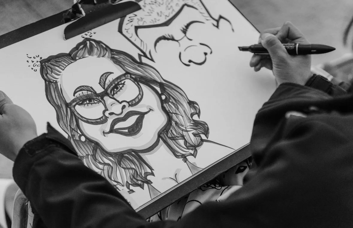 Person drawing a caricature of a woman wearing glasses, with cartoon face sketches nearby.