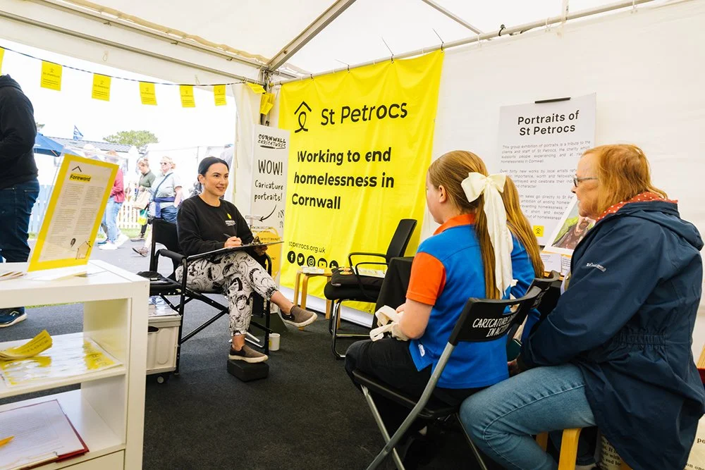 A woman sitting on a chair conducting a caricature portrait at a booth, with two women sitting facing her. The booth displays a yellow banner that reads "St Petroc's Working to end homelessness in Cornwall," and additional informational posters and signs are visible.