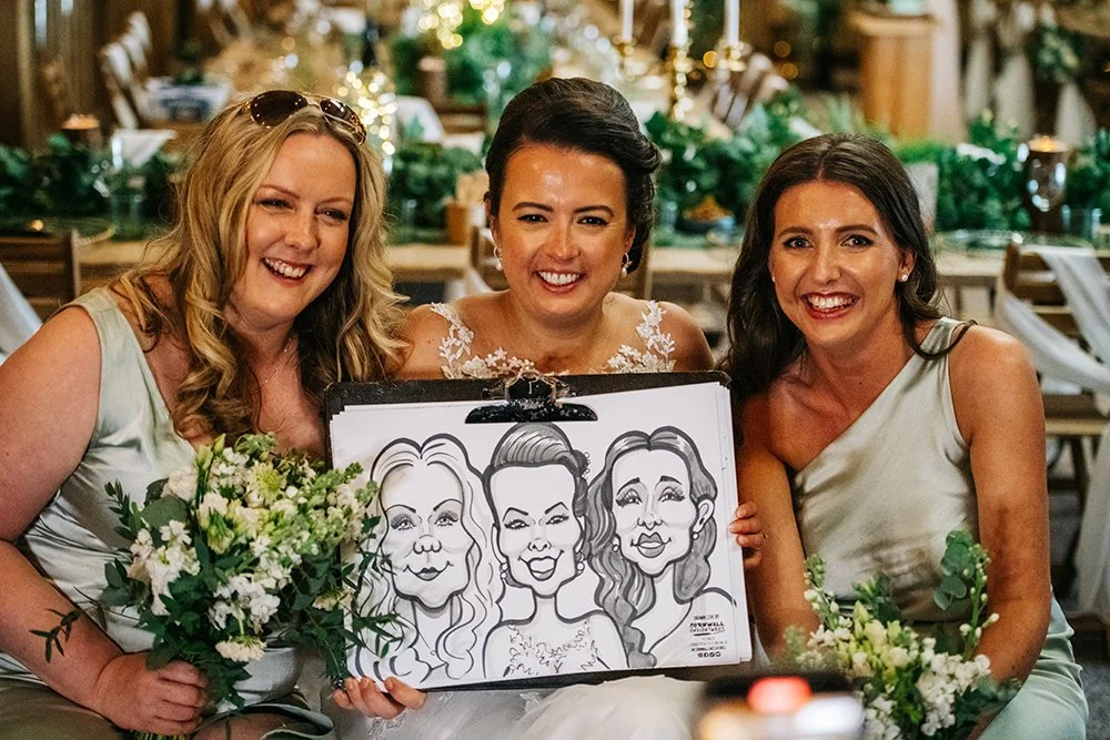 Three women smiling and holding a caricature drawing of themselves at a wedding reception.