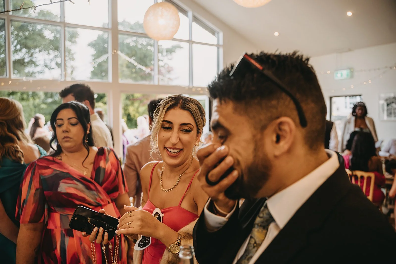 People at a social gathering indoors, diverse group including a woman in a pink dress showing something on her phone and a man in a suit laughing covering his mouth. Large windows, bright natural light, and caricatures being drawn out of the frame.