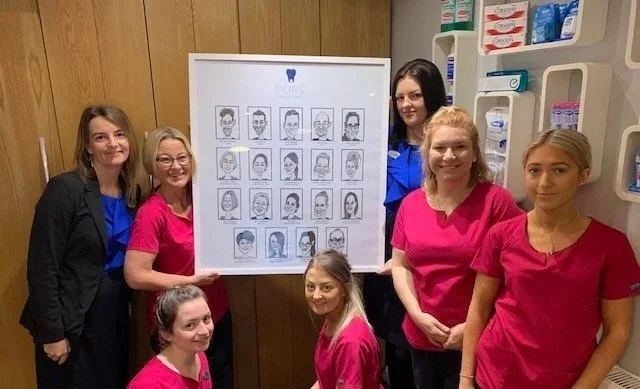 Six women in a dental office, some wearing pink scrubs and one in a blue shirt, standing around a poster with various illustrated smiles, with dental supplies on shelves behind them.