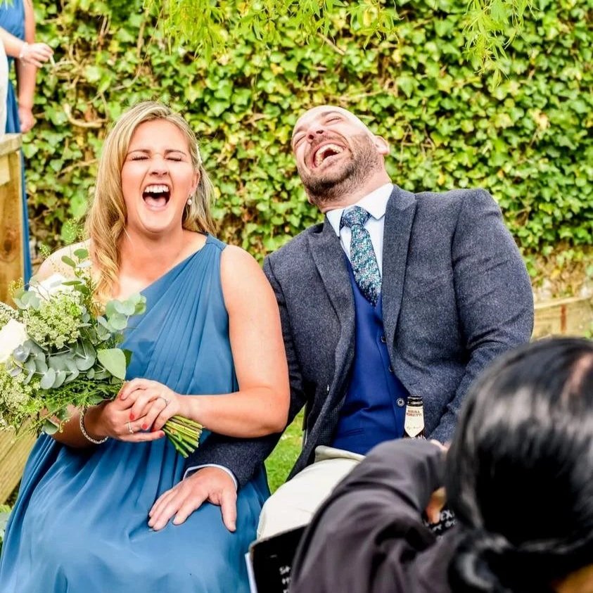 A bridesmaid in a blue dress and a man in a grey suit laughing together at a live caricature drawing at an outdoor wedding drinks reception, while the Caricature artist spins the clipboard around, against a green leafy background.