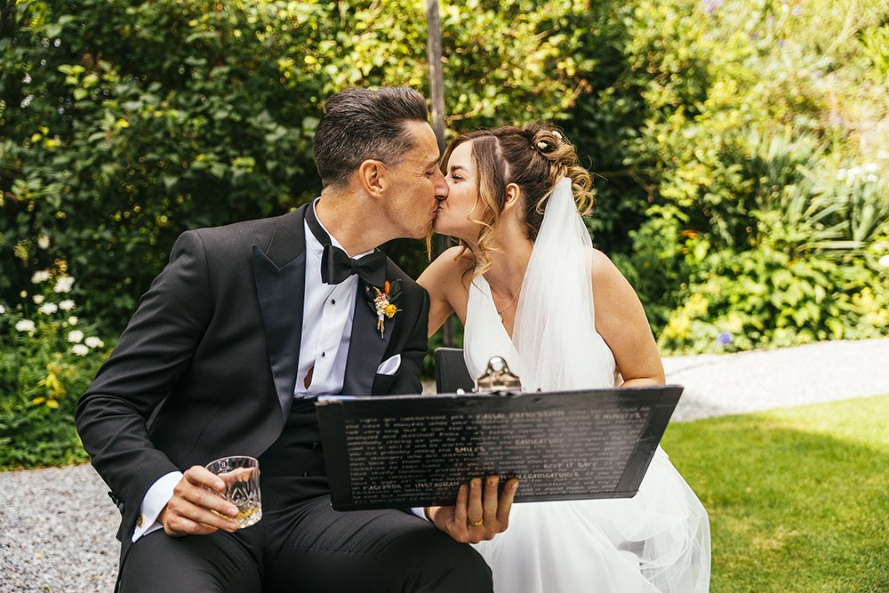 A newlywed couple in wedding attire sharing a kiss outdoors, with a man in a tuxedo and a woman in a white wedding dress and veil, surrounded by greenery.