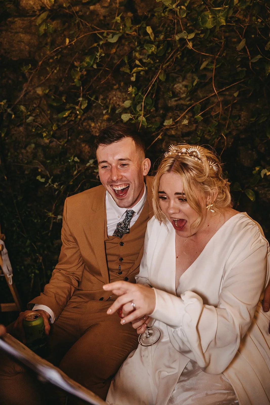 A man in a tan suit and a woman in a white dress celebrating their wedding, sitting together with their mouths open, smiling and holding drinks, in front of a leafy background.