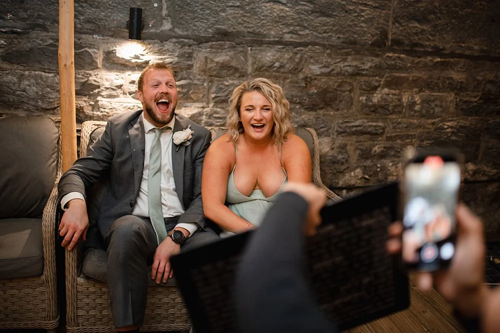 A man and woman sitting on a sofa, laughing and smiling, as a person takes a photo of them with a smartphone.