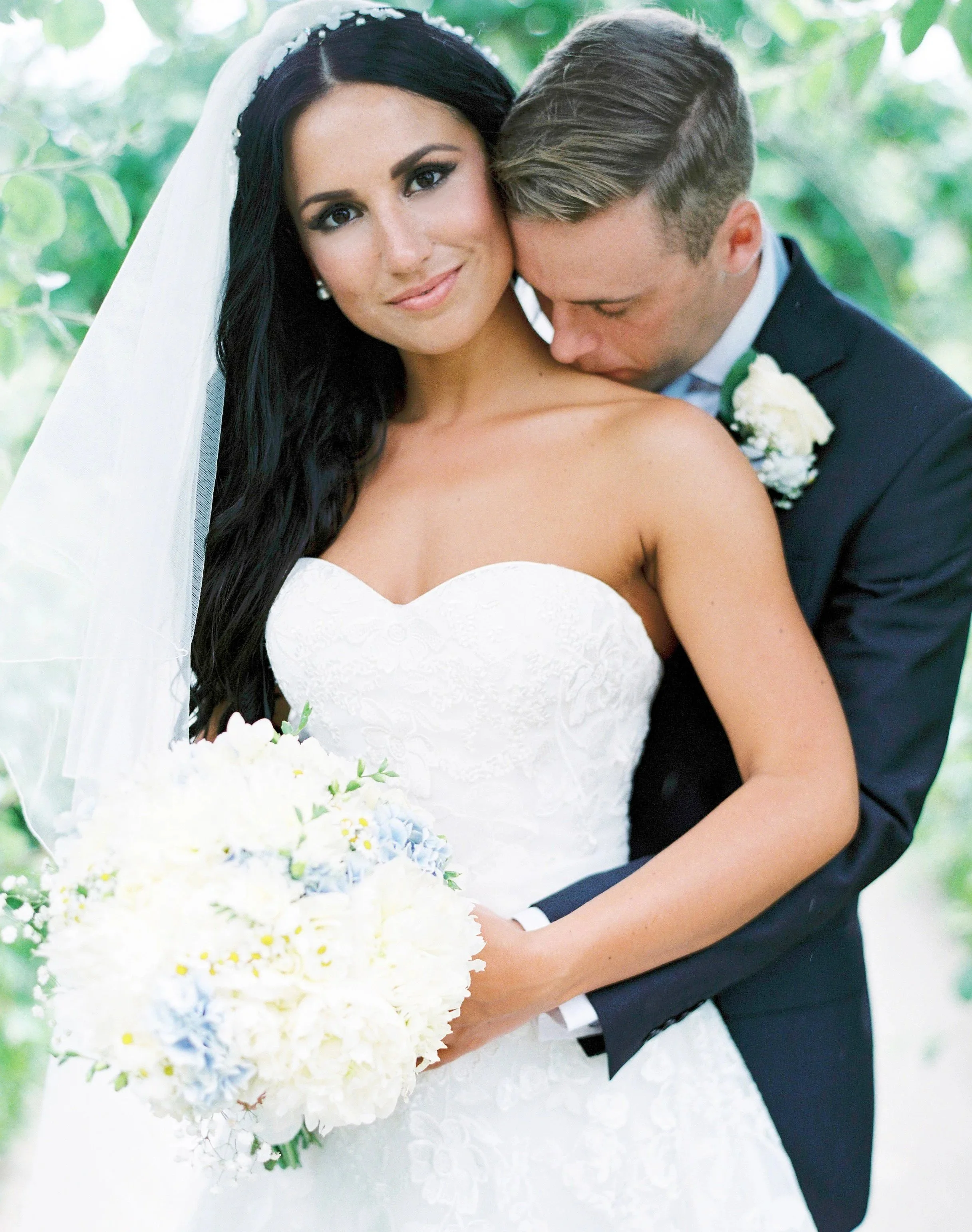 A bride and groom share a tender moment at their wedding, with the groom kissing the bride's neck and the bride smiling softly. The bride has long dark hair with a veil, and the groom is dressed in a dark suit with a white shirt and boutonniere. The background is blurred greenery, giving a natural outdoor setting.