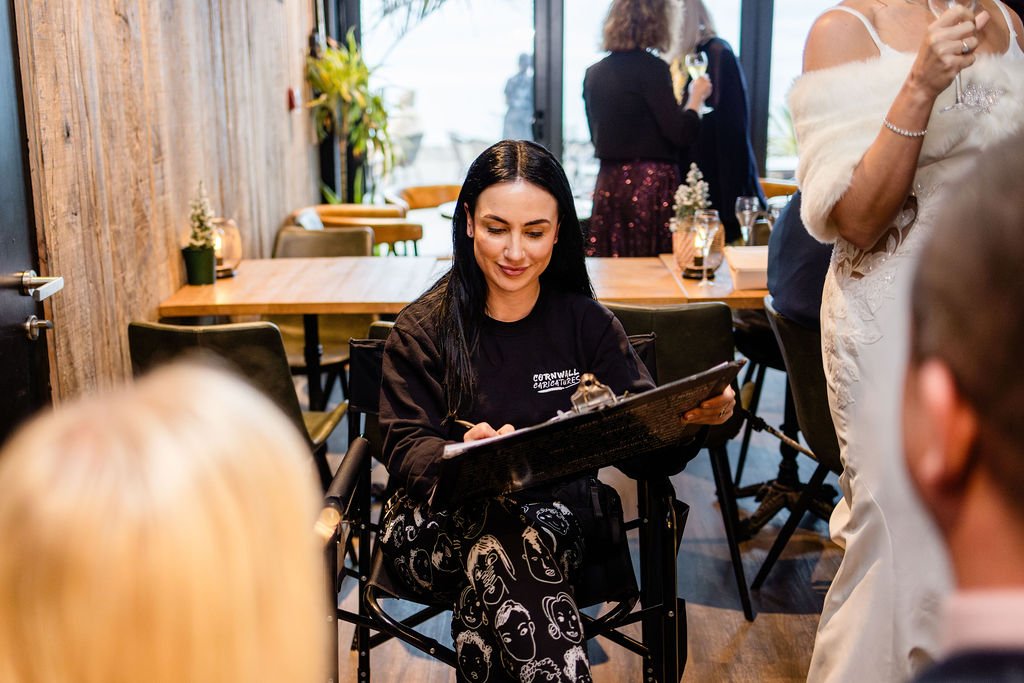A woman with long dark hair, wearing a black sweatshirt and patterned pants, is sitting and drawing. There are other people in the background, some standing and talking, with a wooden wall and large windows behind them.