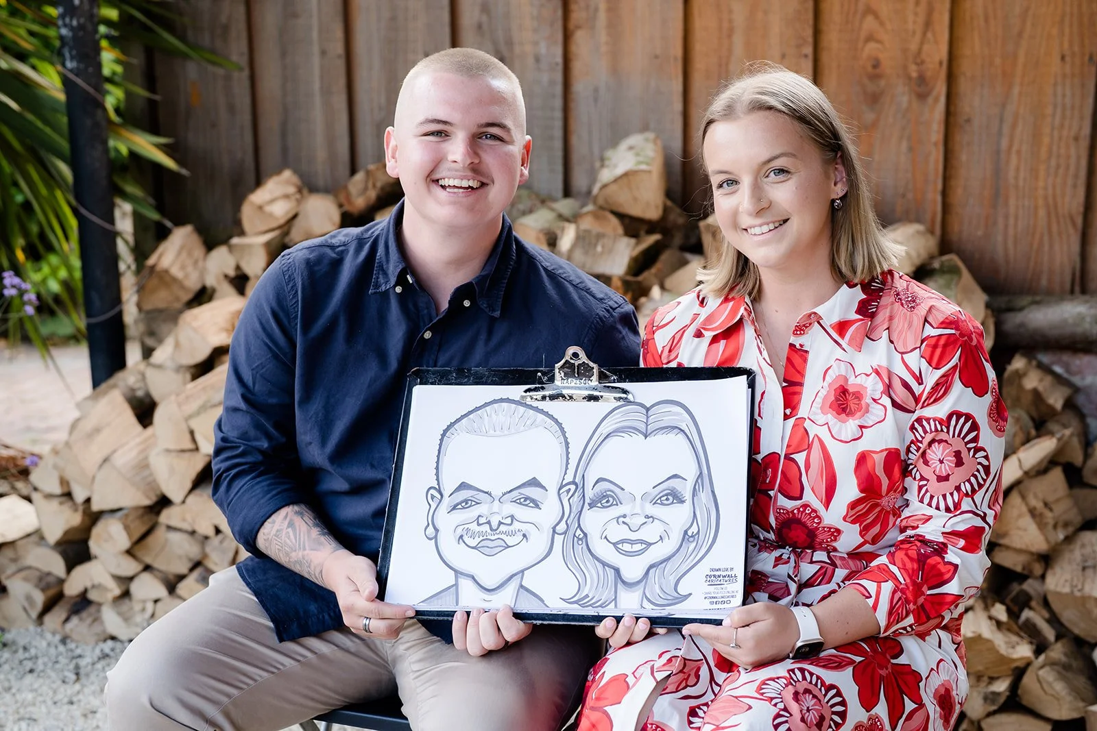 A smiling man and woman sitting outdoors, holding a caricature drawing of themselves on a clipboard, with a woodpile and fence in the background.