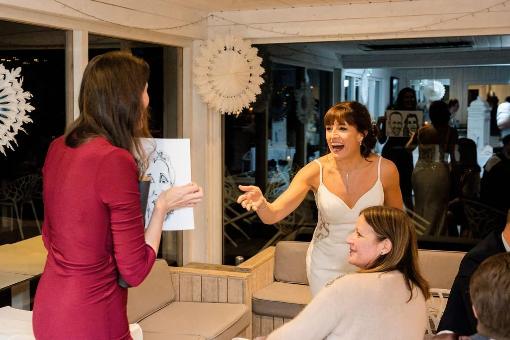 Women at a social gathering, with one woman wearing a white dress excitedly talking to another woman in a red dress, while a third woman in a beige sweater looks on and smiles.