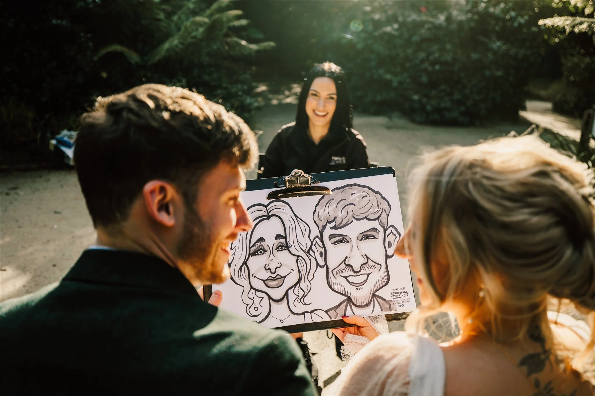 People gathered outdoors, listening to a woman speaking with a caricature drawing of a man and woman in front of her.