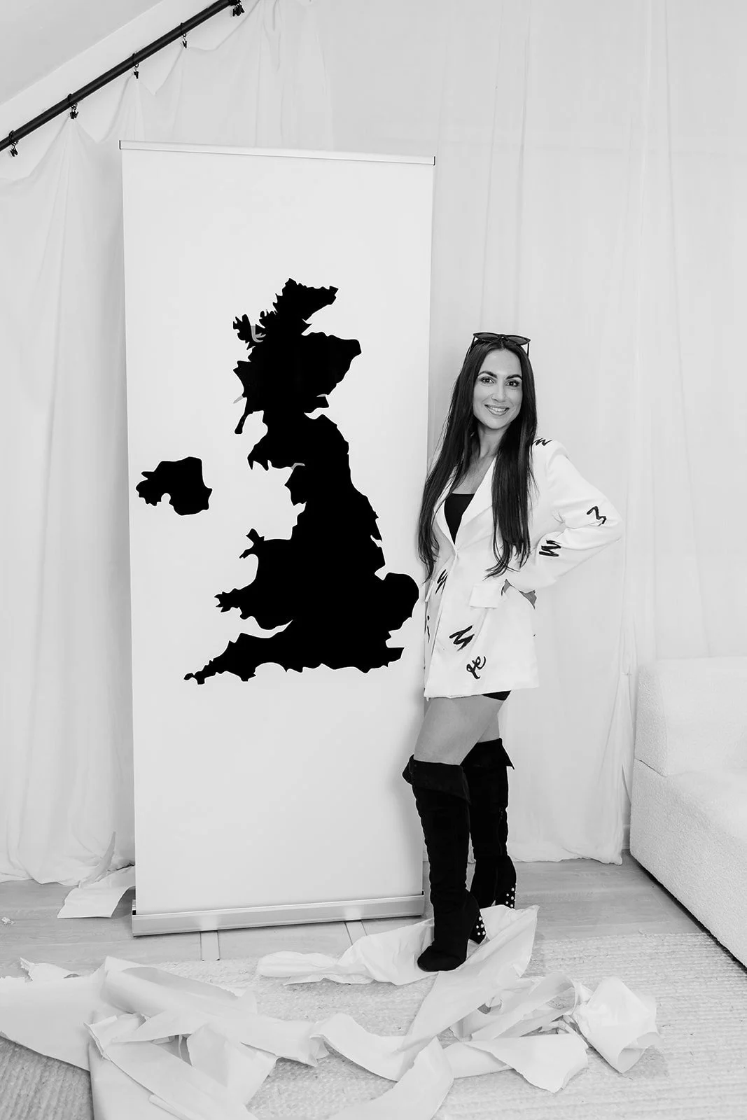 A woman standing next to a large black silhouette map of the United Kingdom on a white backdrop, smiling and posing with one hand on her hip.