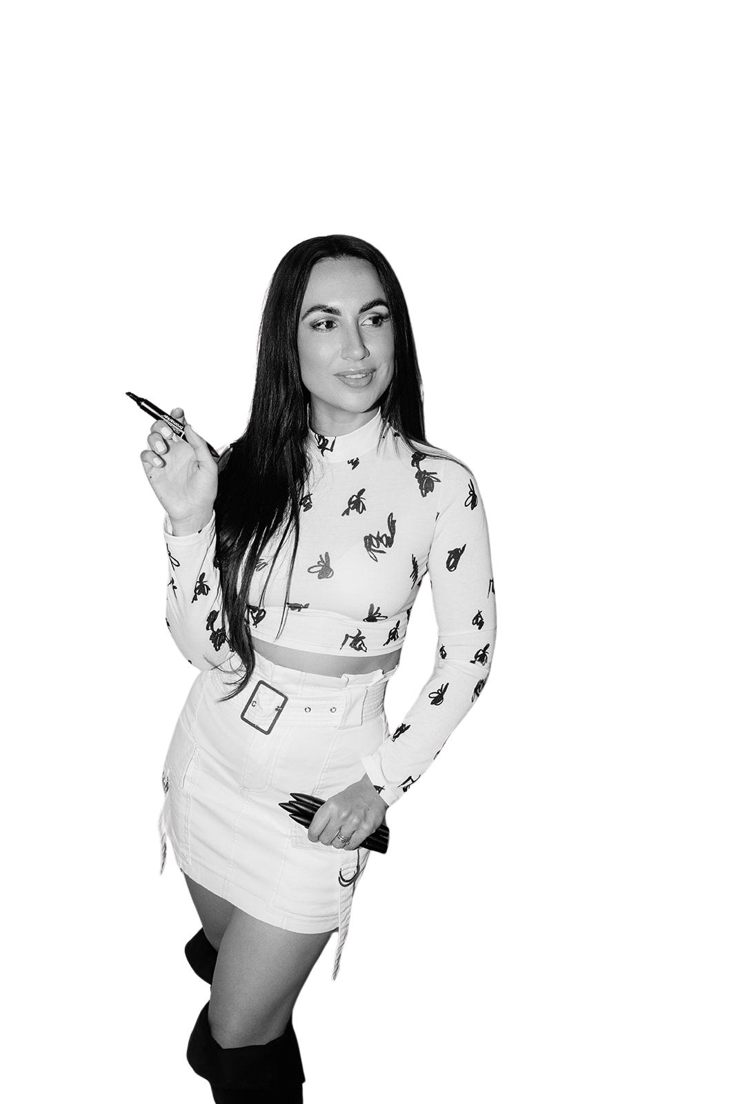 Black and white photo of a young woman with long dark hair, holding a pen in her right hand and a small bundle of pens in her left hand, standing against a white background. She is wearing a long-sleeved, patterned crop top and a high-waisted skirt.
