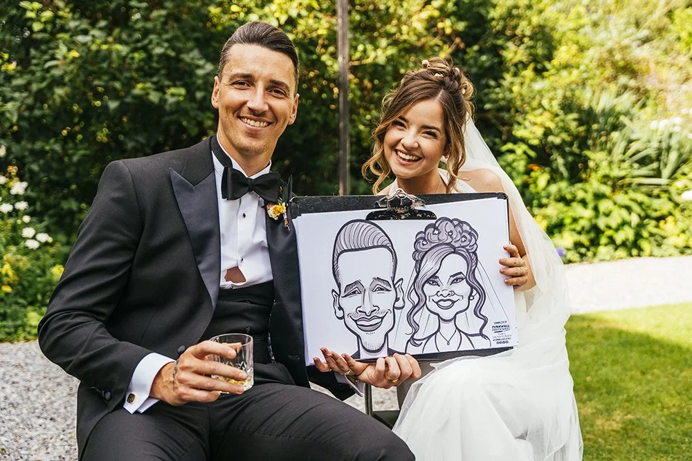 Smiling bride and groom in wedding attire holding a caricature drawing of themselves outdoors