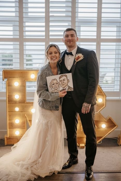 Bride and groom standing together at their wedding, holding a caricature portrait of themselves, with illuminated marquee letters in the background.