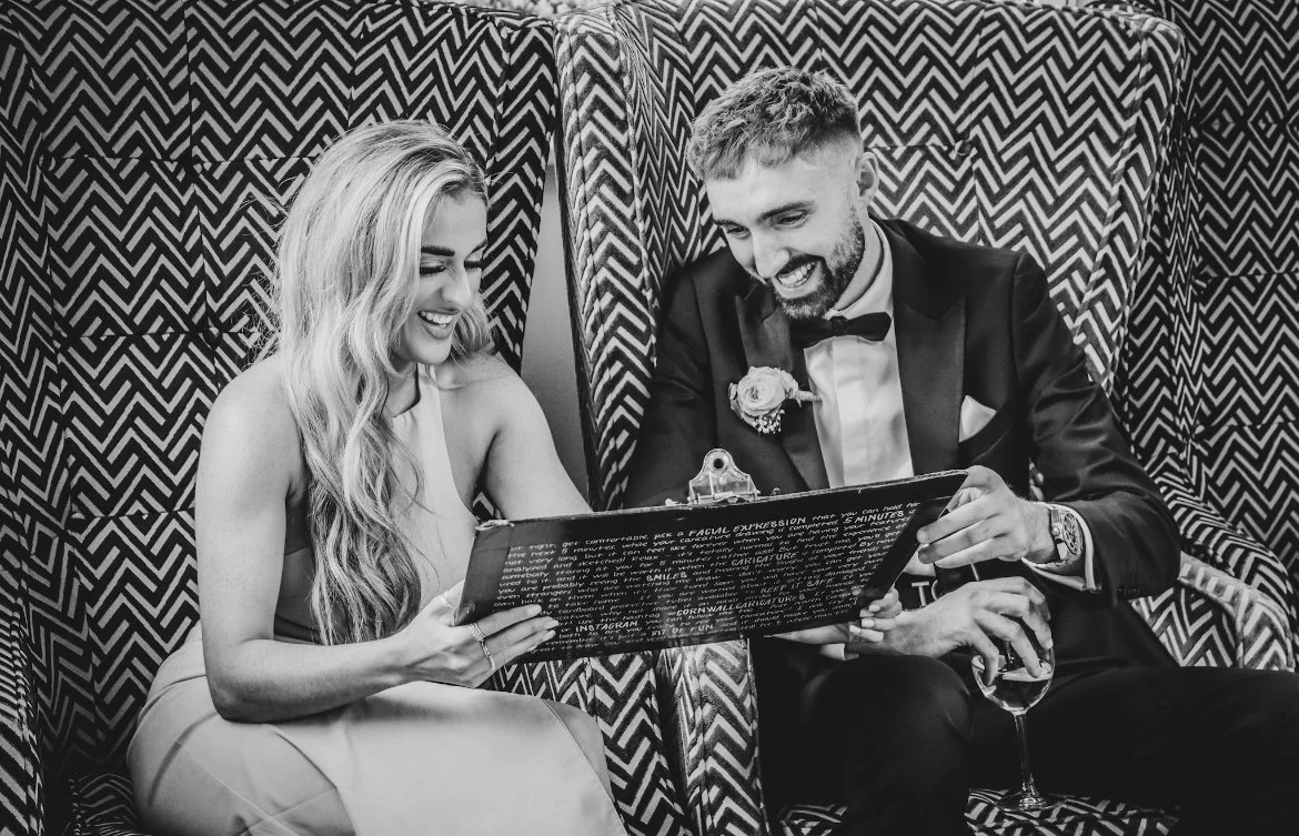 A man in a tuxedo and a woman in a dress sitting together, smiling and looking at artwork, with patterned chairs behind them.