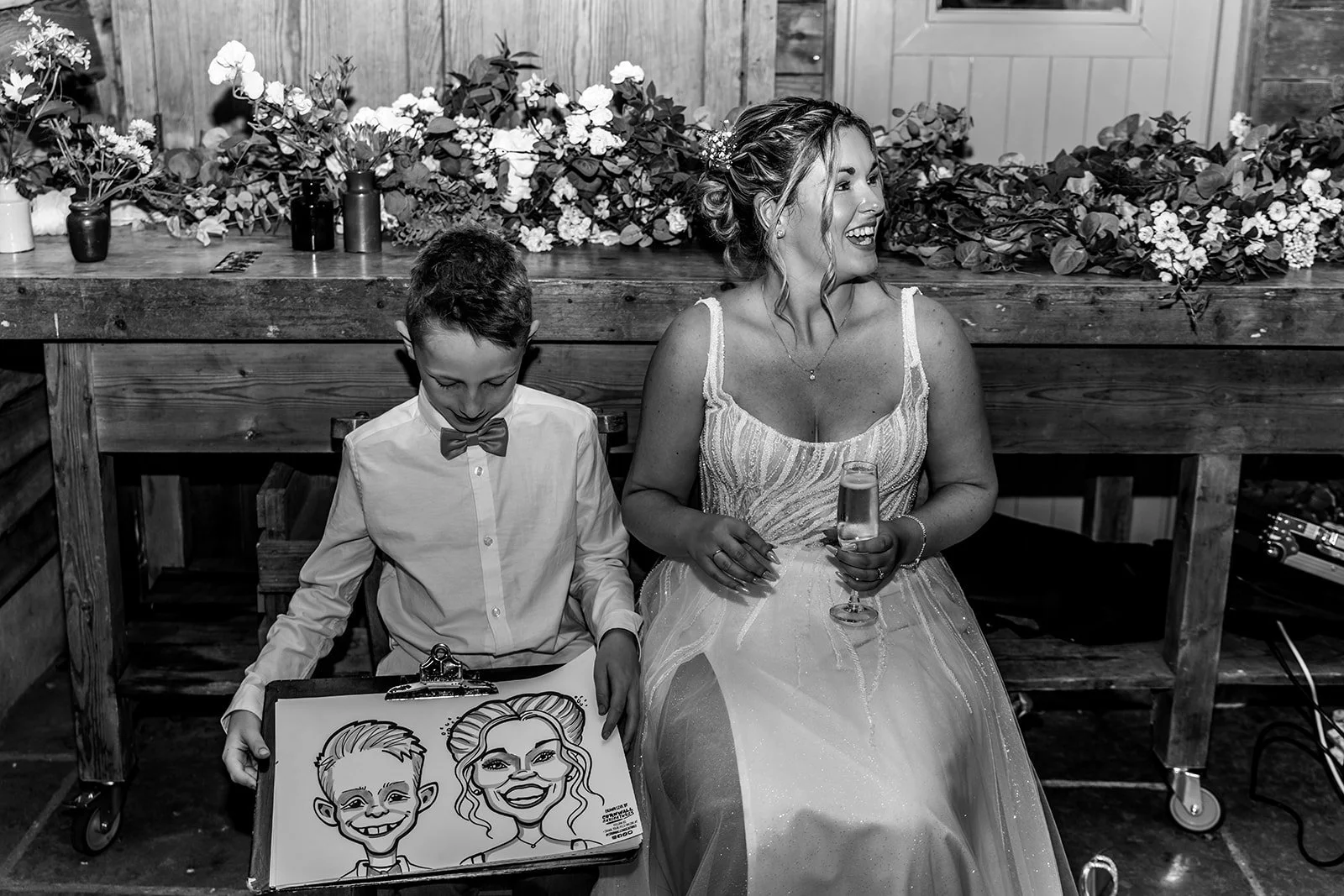A woman in a wedding dress sitting on a bench, holding a glass of champagne, smiling and looking to her right, next to a young boy in a white shirt and bow tie, sitting on the same bench and looking down at a caricature drawing of themselves.