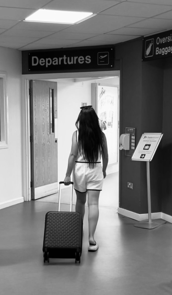 A woman with long dark hair wearing a sleeveless white dress walking through an airport towards the departure area, pulling a small rolling suitcase behind her.