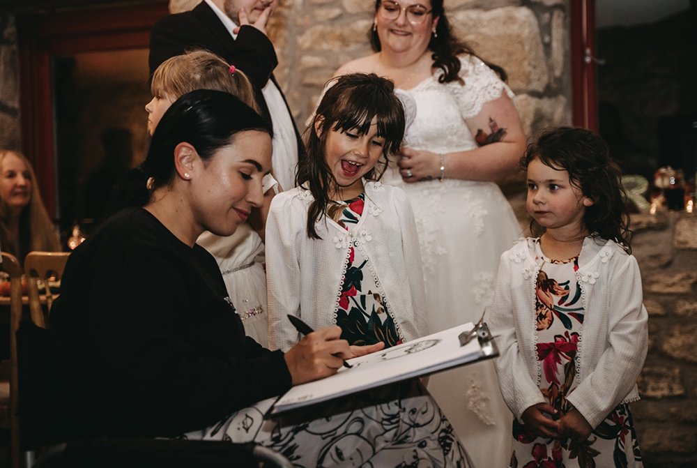A woman is drawing caricatures while three young girls and a woman in a white dress watch, smiling.