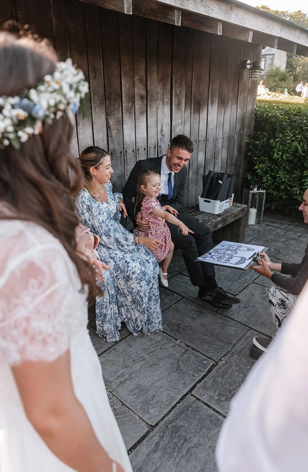 Group of people at a wedding, with a woman in a white dress and floral crown partially visible, a woman in a blue patterned dress, a young girl in a floral dress, and a man in a suit, all smiling and pointing at a drawing in a sketchbook held by a wo