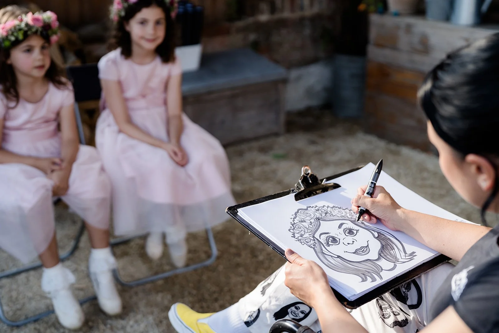 An artist drawing a caricature of two young girls in pink dresses, one wearing a flower crown, as they sit for their portraits outdoors.