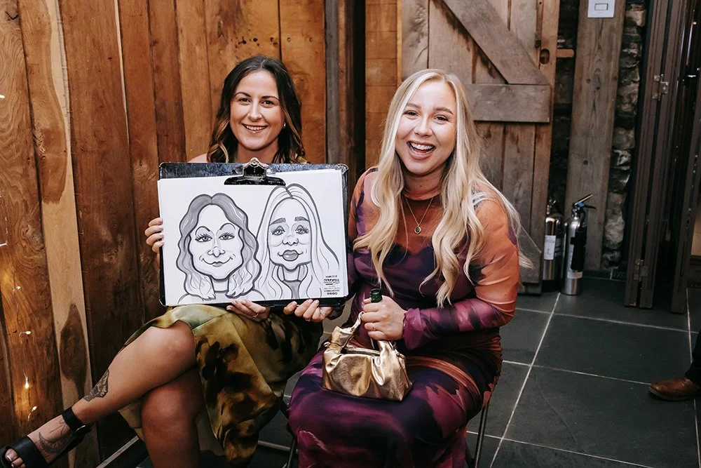 Two women sitting on chairs, one holding a caricature drawing of two women, in a wooden indoor setting.