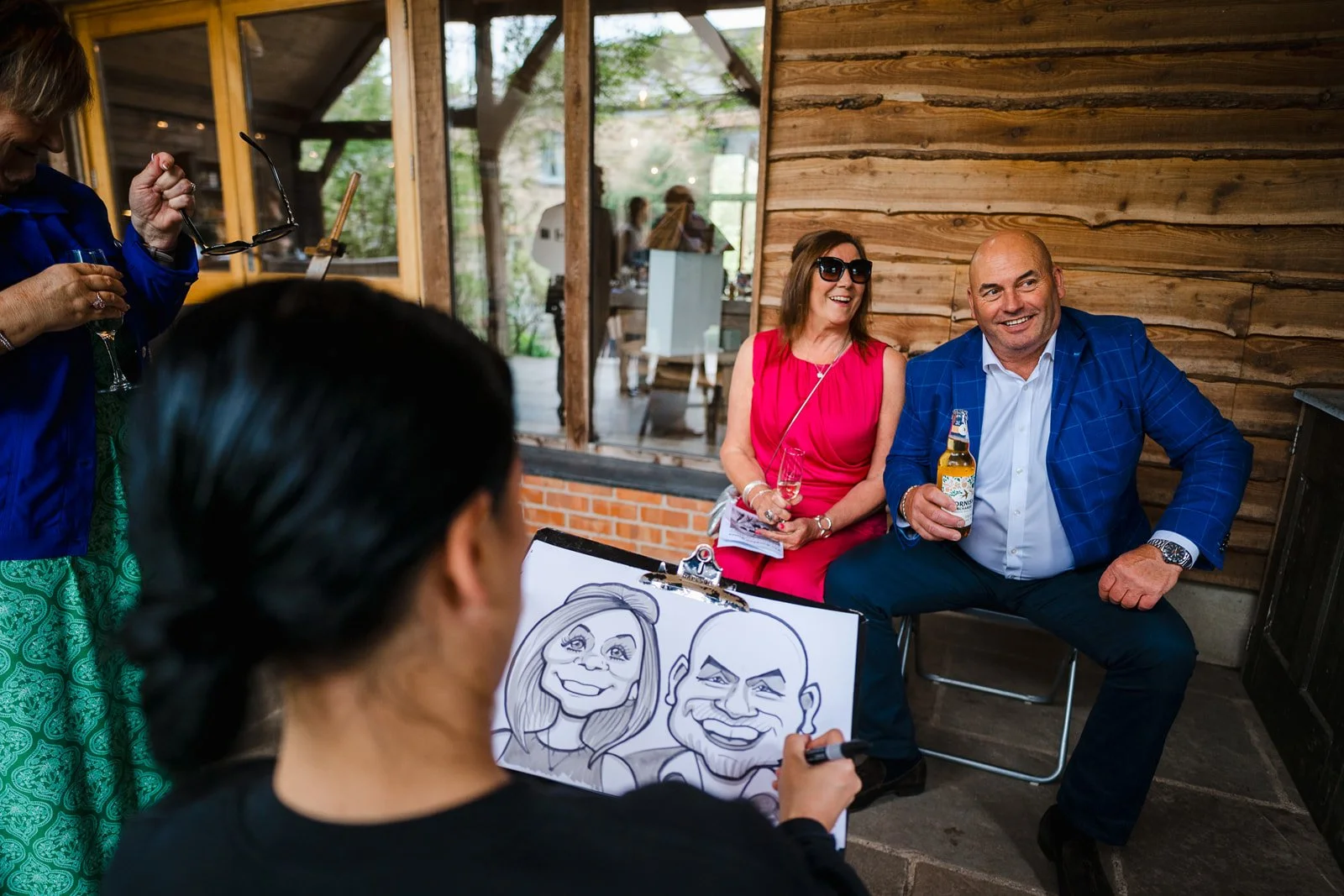 A woman is drawing caricatures of two smiling people at a social event held indoors with wooden walls and large windows, with two people sitting and posing for the drawing.