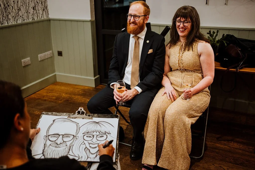 A woman is drawing a caricature of a man and woman seated on chairs, both holding glasses, at an indoor event.