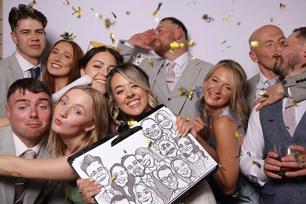 Group of people celebrating, posing with caricatures, confetti in the air, some smiling and others kissing, at a party or wedding.