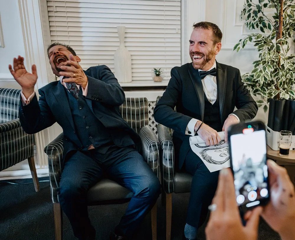 Two men in tuxedos sitting in a living room, laughing and sharing a joyful moment while one person takes a photo of them.