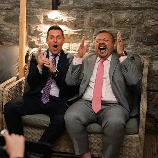 A male couple in wedding suits sitting on a wicker bench, laughing and reacting with surprise and excitement to their caricature, with a stone wall backdrop.