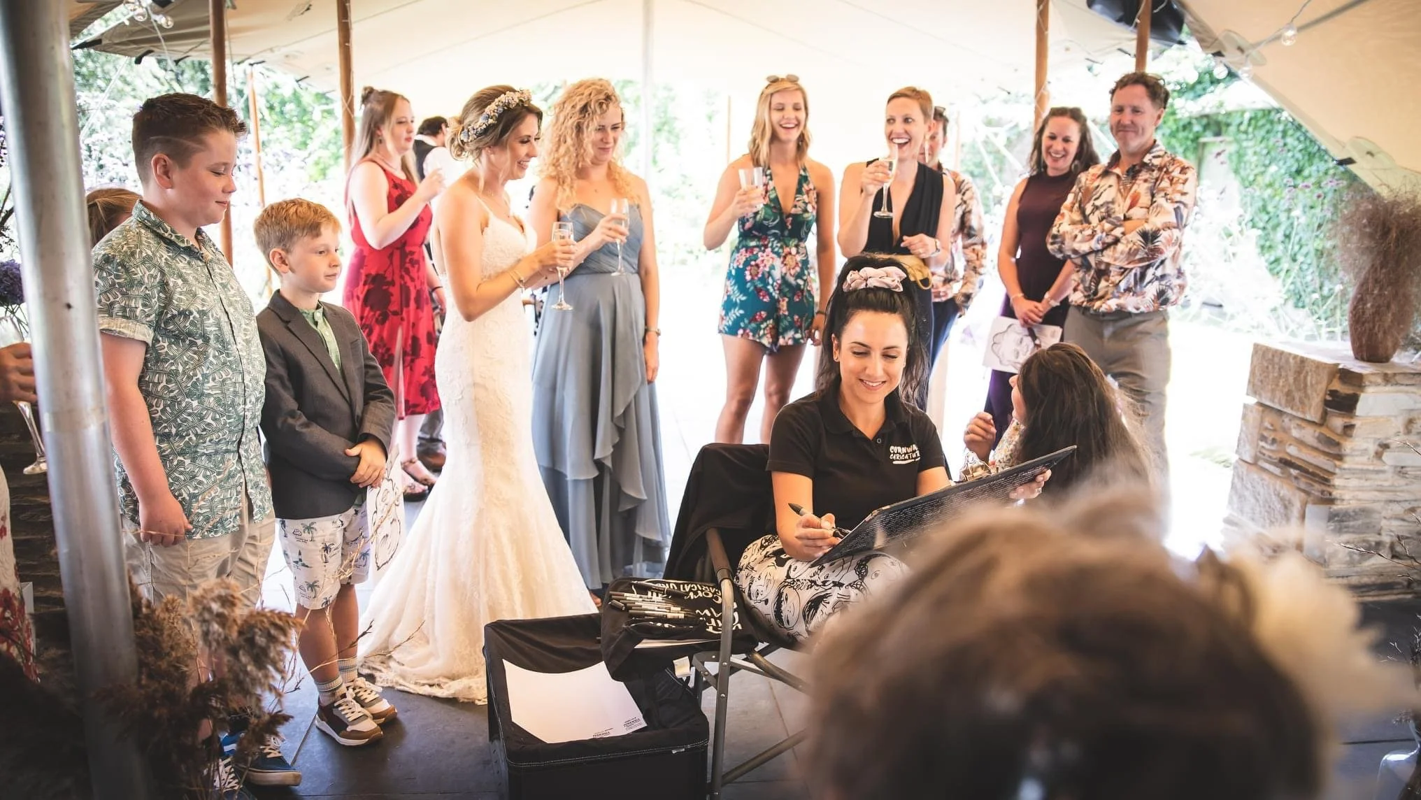 Group of people at a wedding celebration, with women wearing dresses and a bride in a white gown, two young boys in front, and a person seated at a table drawing guests.