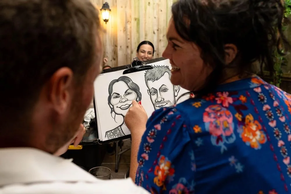 A woman drawing caricatures of two people on a sketchpad as others watch at a social gathering.