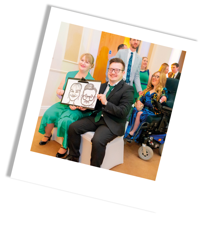 Group of people at an event, with one person holding a drawing of faces, others smiling and dressed in formal attire, including a woman in a green dress and a woman in a wheelchair.
