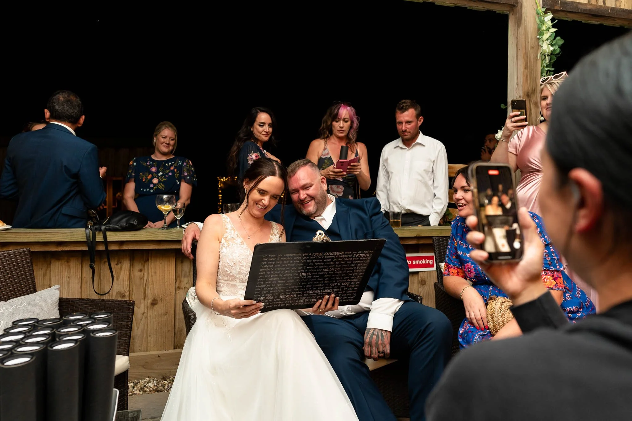 At a wedding reception, a bride and groom sit together smiling at their portrait illustration, surrounded by guests taking photos and mingling, with lively music and drinks on the table.