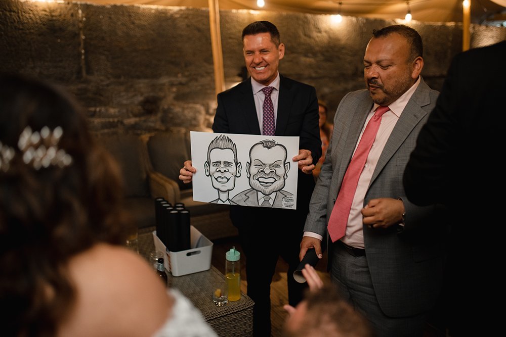 Man in dark suit holding caricature drawing of two men at a celebration or party, with smiling guests around.