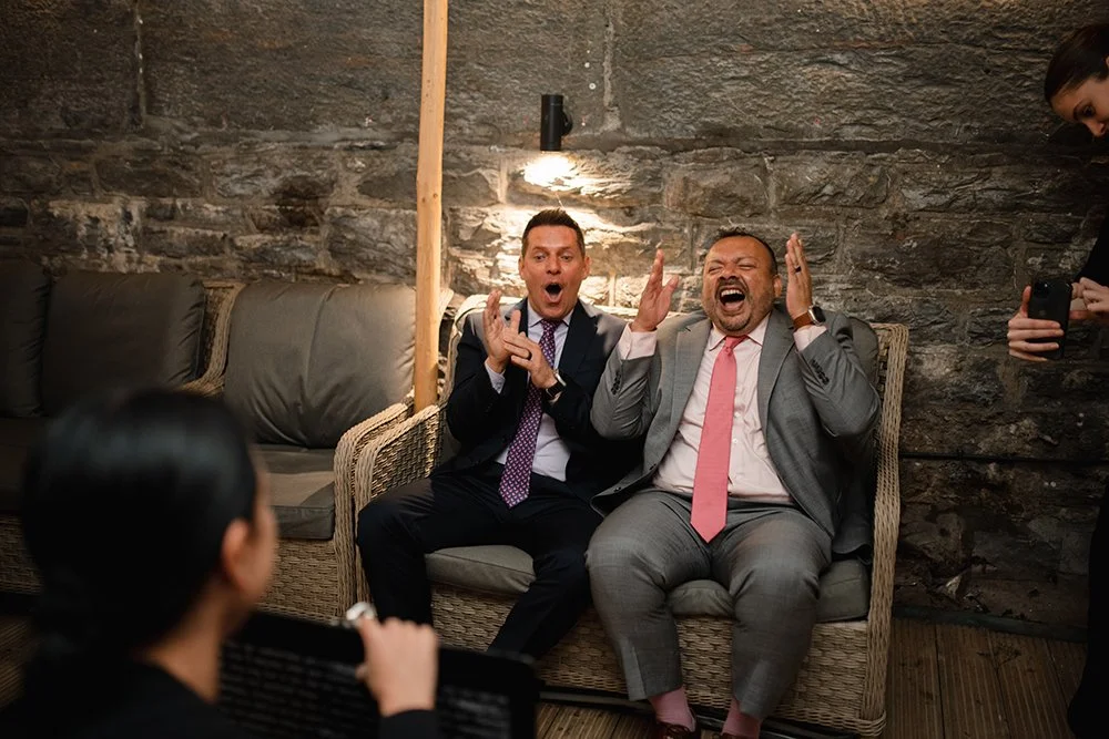 Two men in suits sitting on a wicker couch, laughing and reacting emotionally in a cozy, rustic setting with stone walls, as a woman in black presents their caricature, and a woman on the right takes a photo.