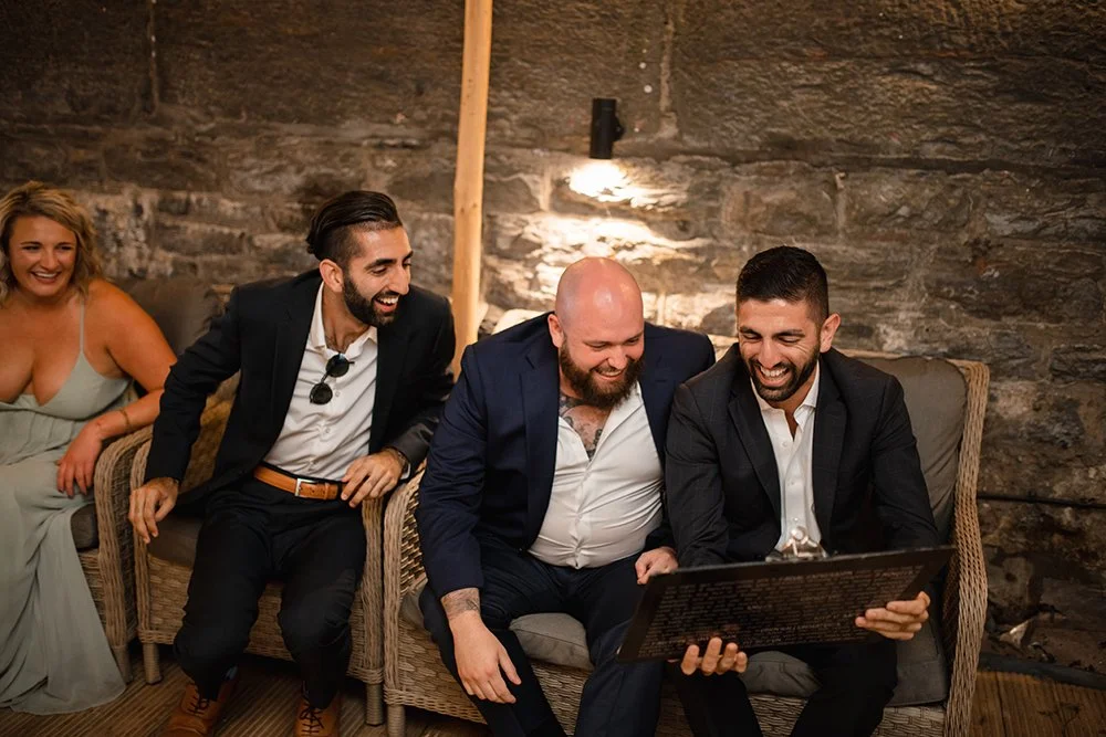 A group of four friends sitting on a wicker sofa in a stone-walled room, laughing and looking at a caricature drawing.