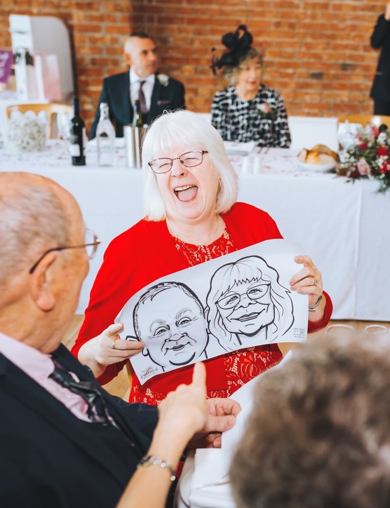 A joyful elderly woman holding a caricature drawing of herself and another person at a social gathering, likely a wedding reception, with people sitting at a decorated table in the background.