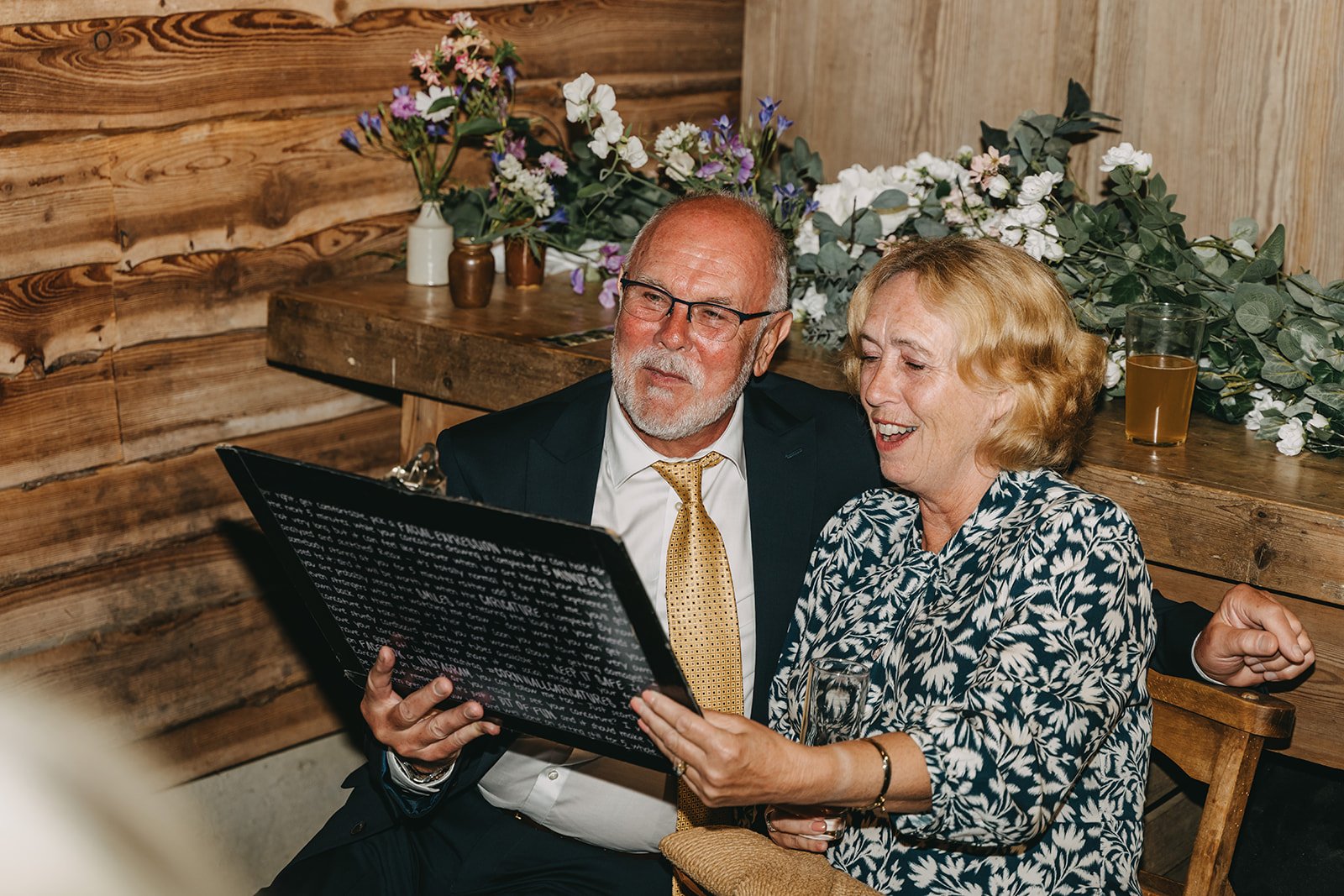 An older man and woman sitting together at a table, looking at a caricature the woman is holding, with flowers and wooden wall in the background.