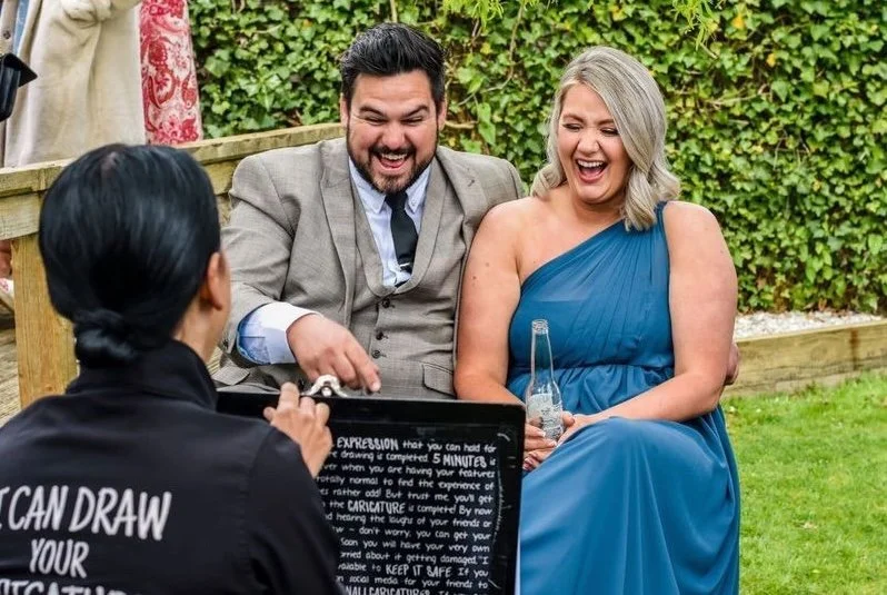 A man and woman laughing and smiling while at an outdoor event, with a woman in black in front of them taking their picture, holding a digital camera. The woman in black is wearing a shirt with text that says, 'I CAN DRAW YOUR CARICATURE'.