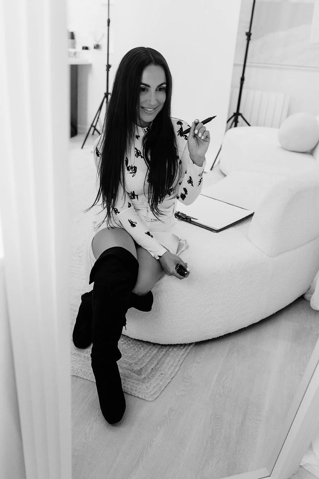 A woman with long dark hair wearing a scribble print top and over-the-knee boots sitting on a sofa, smiling, holding a marker, in a photography studio with lighting equipment in the background.