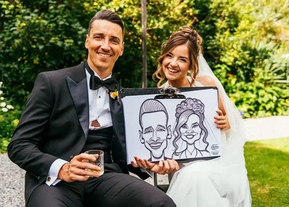 A happy bride and groom in wedding attire sitting outdoors, holding a caricature sketch of themselves on a clipboard. The groom is in a tuxedo, and the bride is in a wedding dress, both smiling.