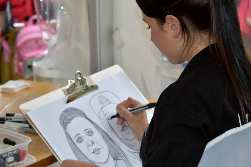 A woman drawing a portrait of a woman with straight hair on a sketchpad, seated at a desk with art supplies.