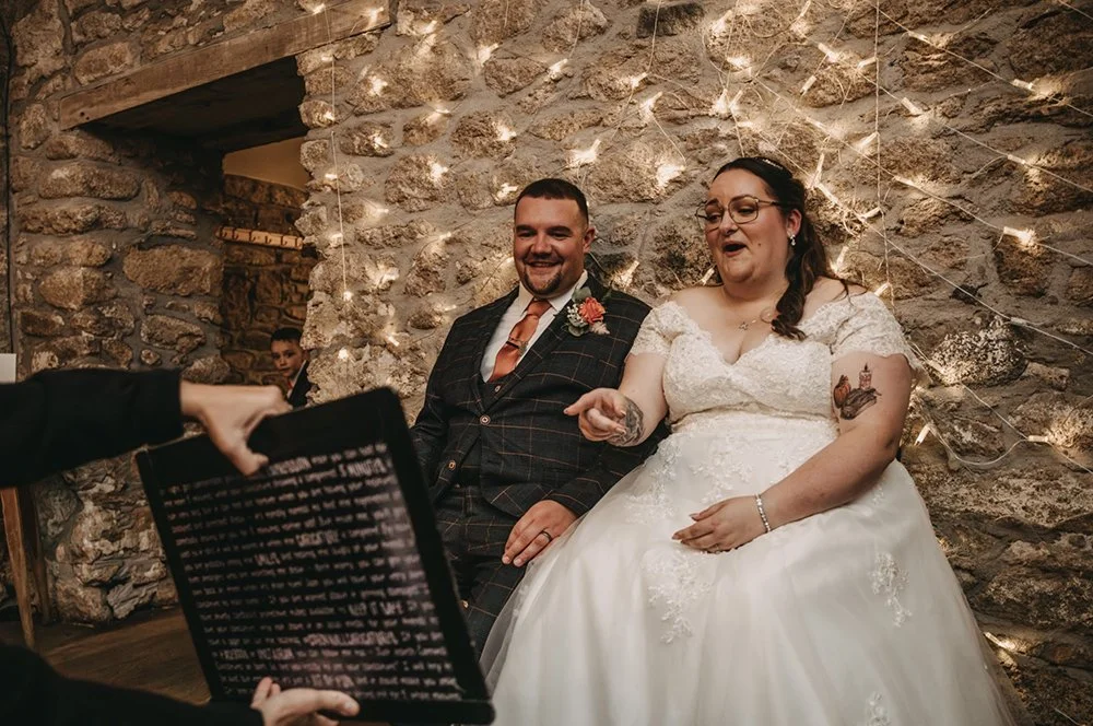 A bride and groom sitting together during their wedding ceremony, smiling, with a stone wall and string lights in the background.