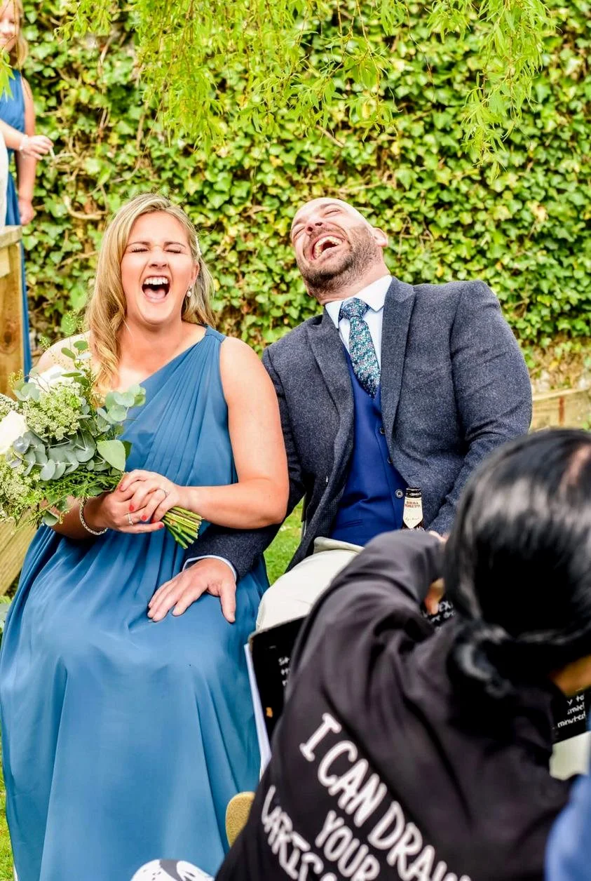 A woman in a blue dress and a man in a suit laughing together at an outdoor event, with a woman holding a bouquet of flowers next to them and a person in the foreground wearing a black hoodie.