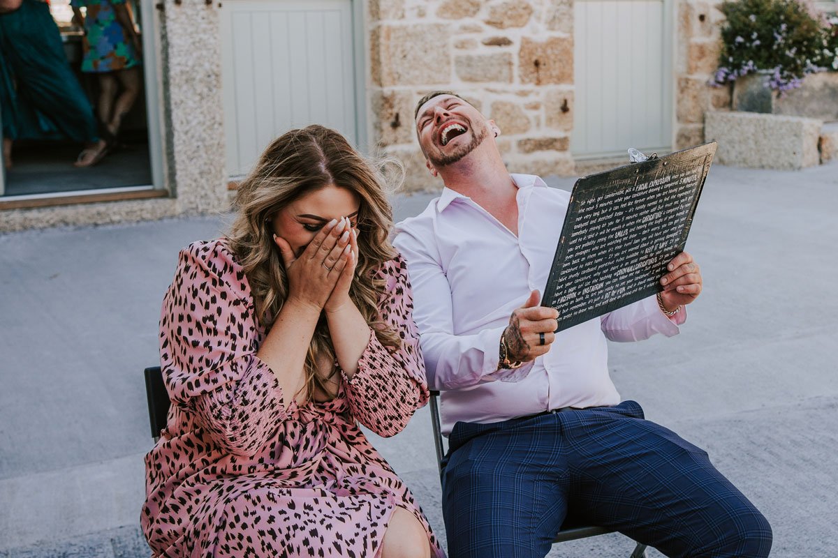 A woman in a pink animal print dress is covering her face and laughing, while a man in a white shirt and blue trousers is sitting beside her, holding a caricature and laughing, outside a stone building.