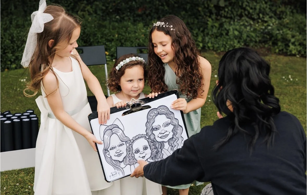 Three young girls in dresses and headbands receiving a caricature drawing from an artist at an outdoor event.