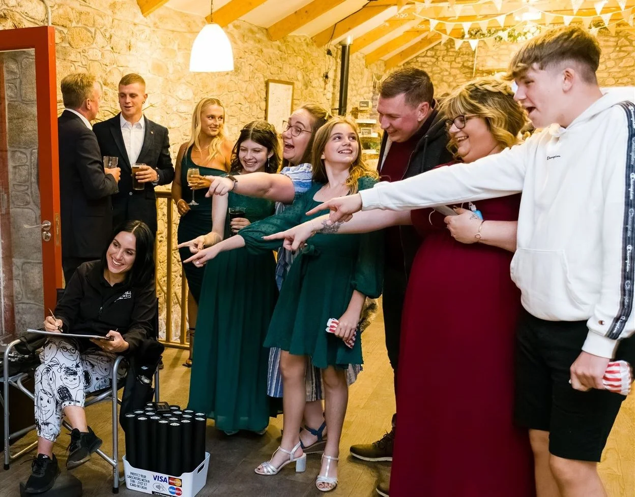 Group of people at a social gathering pointing and smiling, with some holding drinks, inside a warmly lit room with stone walls and a sloped wooden ceiling.